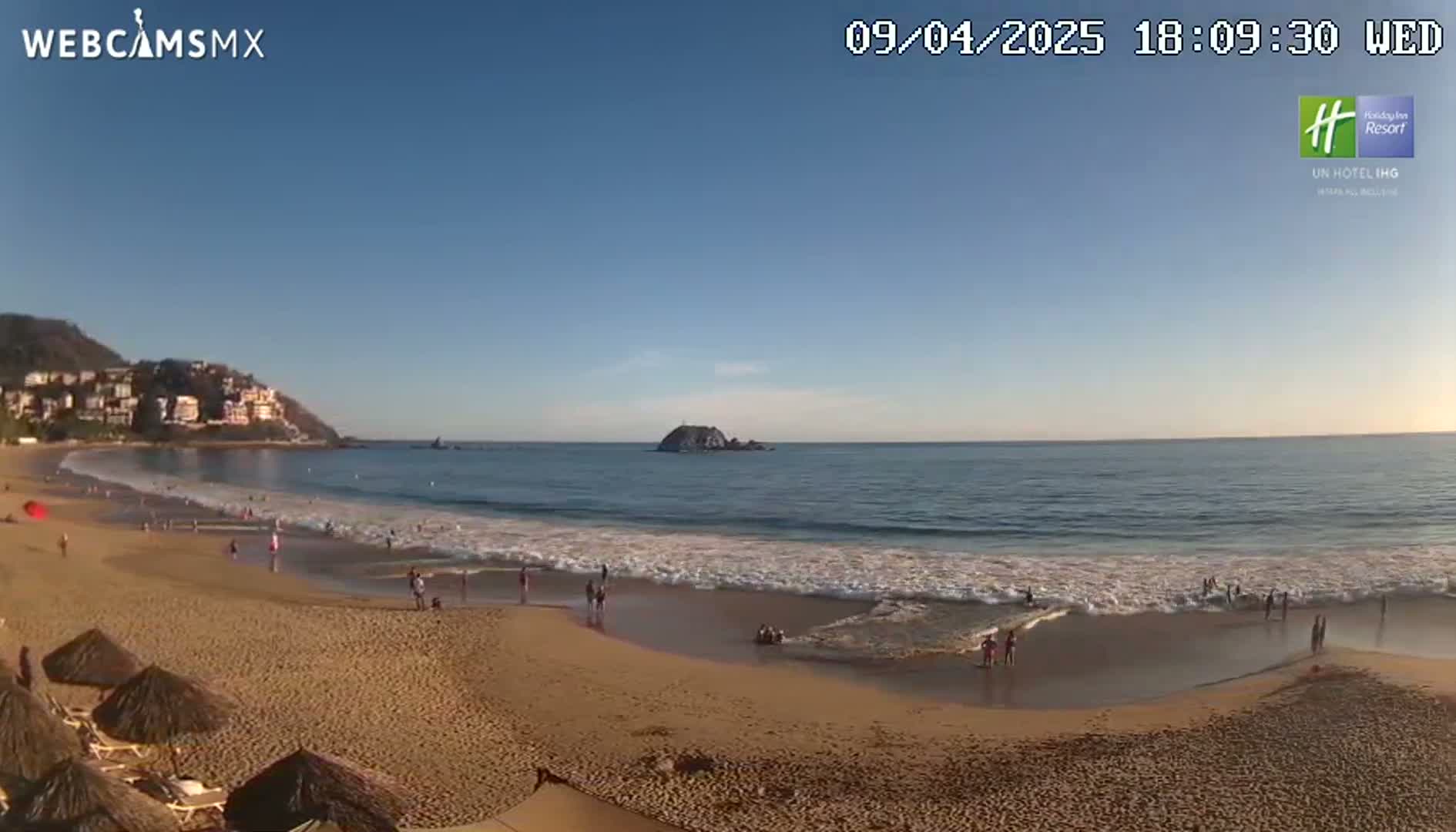 A sandy beach with small waves and a few people on it is next to a calm ocean with a small island in the distance, all under a clear blue sky.