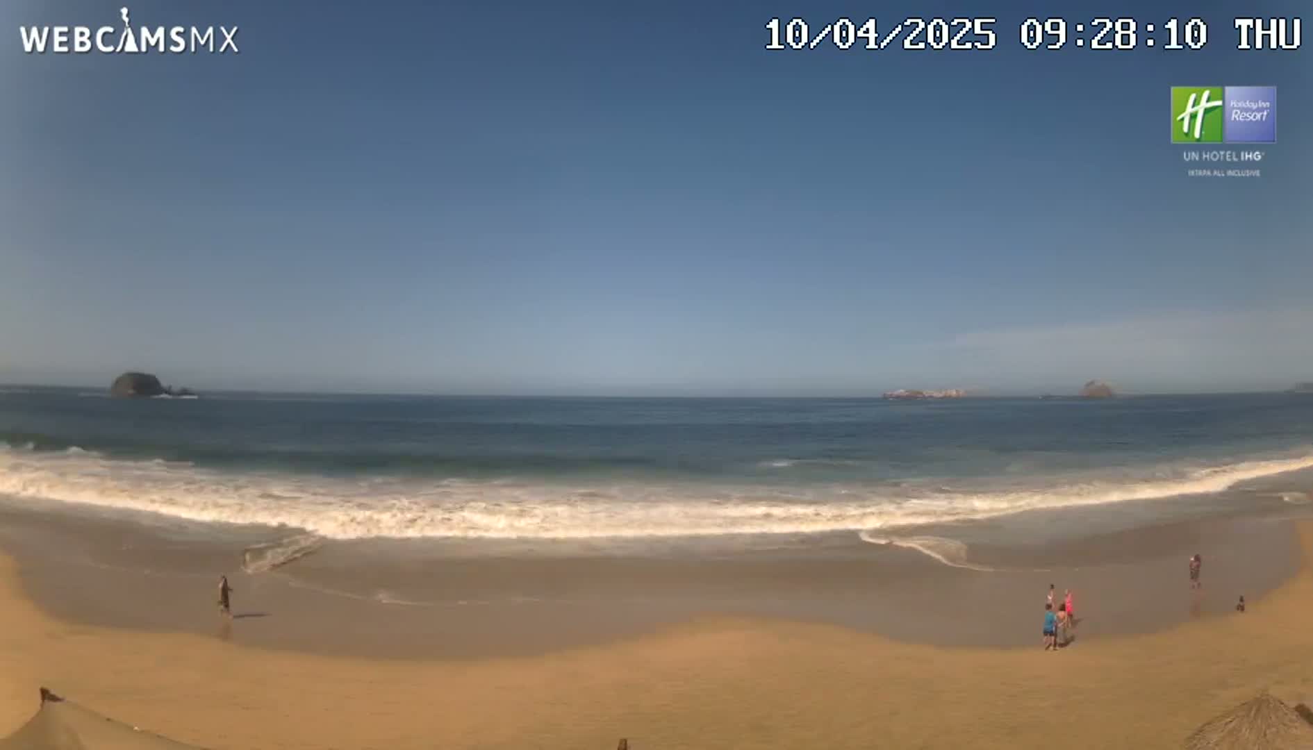 A sandy beach with gentle waves under a clear blue sky shows several people near the water's edge.