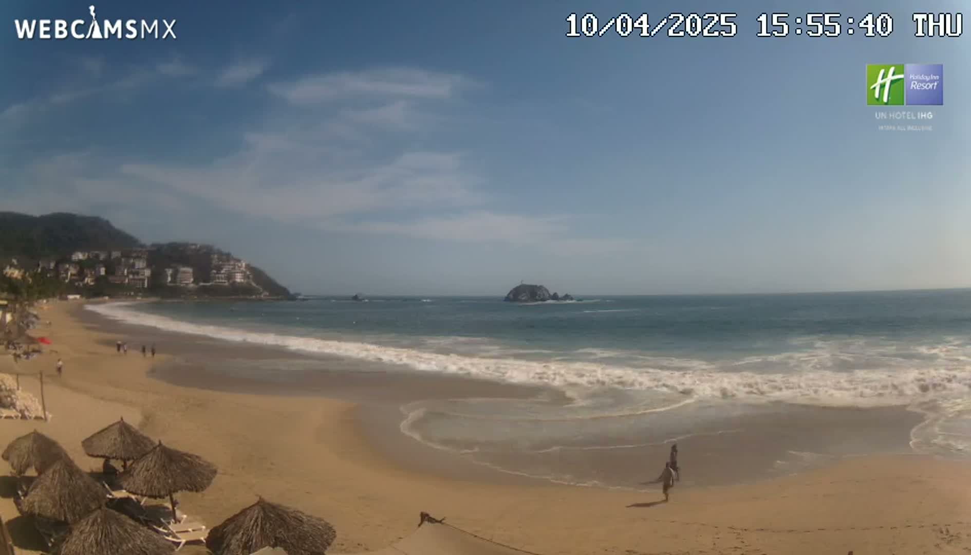 A sandy beach with gentle waves laps the shore under a mostly sunny sky, with buildings and hills visible in the distance.
