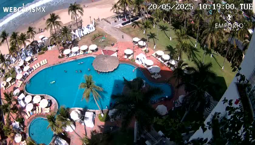An aerial view shows a resort pool with many people swimming and lounging around it, surrounded by palm trees and a beach in the background on a sunny day.