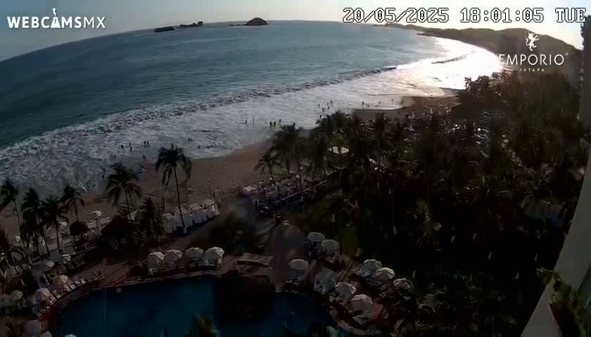 An ocean beach with waves breaking on the shore, palm trees, a resort pool, and some people are visible at sunset.