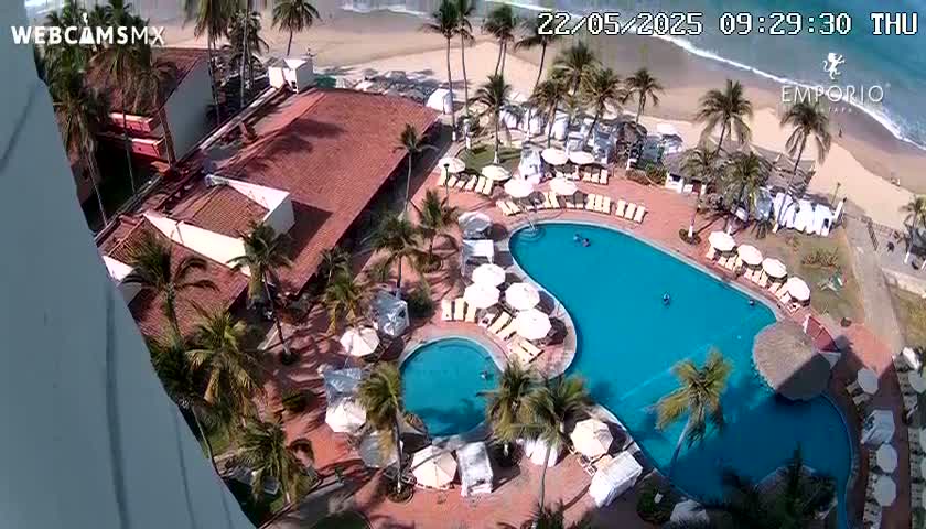 An aerial view shows a resort with a large, kidney-shaped swimming pool and surrounding lounge chairs, palm trees, and buildings, all situated next to a beach with gentle waves on a sunny day.