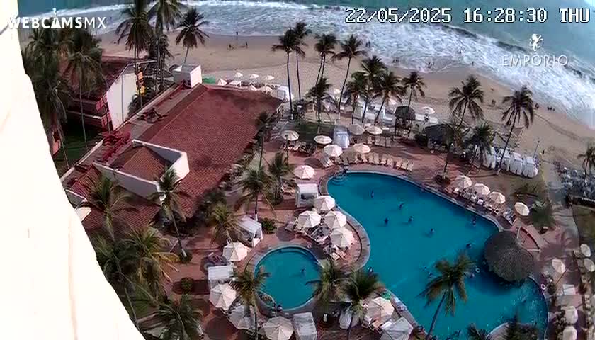 An aerial view shows a resort pool area with palm trees, a beach with waves, and a partially visible building under a partly sunny sky.