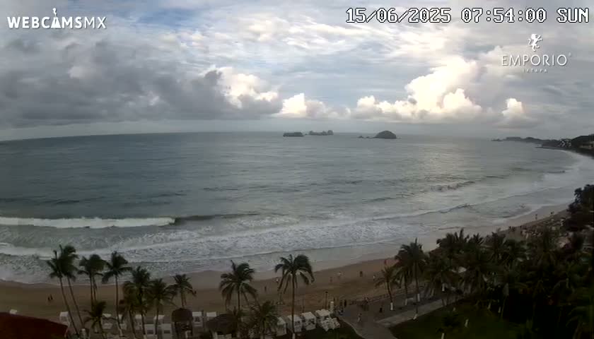 A partly cloudy sky overlooks a beach with palm trees, gentle waves, and distant islands.