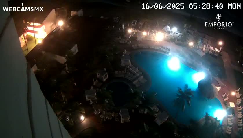An illuminated swimming pool at night is surrounded by buildings and palm trees.