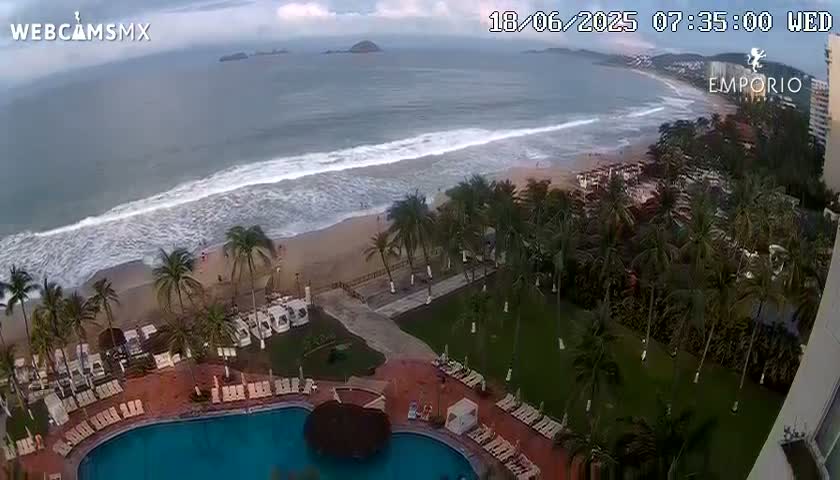 An aerial view shows a resort pool and surrounding palm trees overlooking a beach with waves rolling in under an overcast sky.