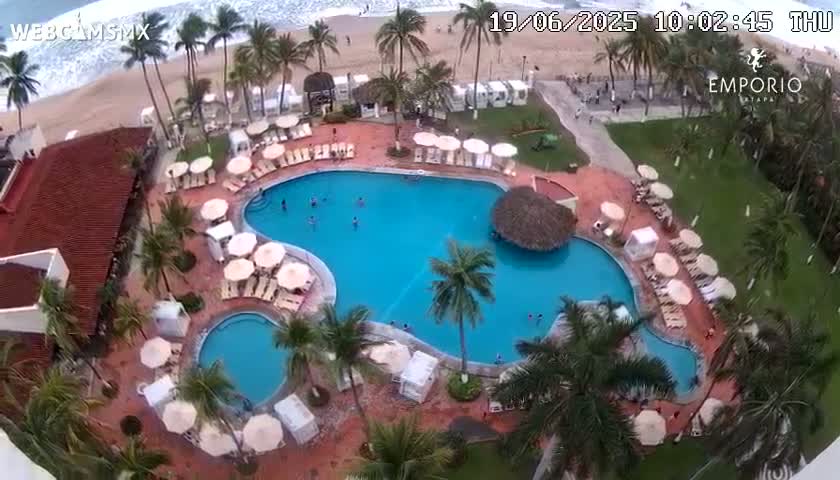 An aerial view of a resort pool area with palm trees, beach umbrellas, and a sandy beach visible in the background under partly cloudy skies.
