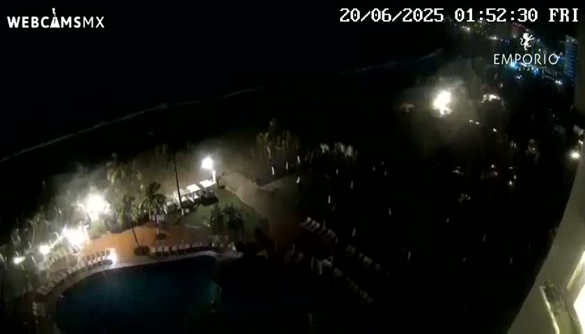 A nighttime, high-angle view shows a resort area with a swimming pool and surrounding buildings and landscaping during a storm with strong winds and rain.