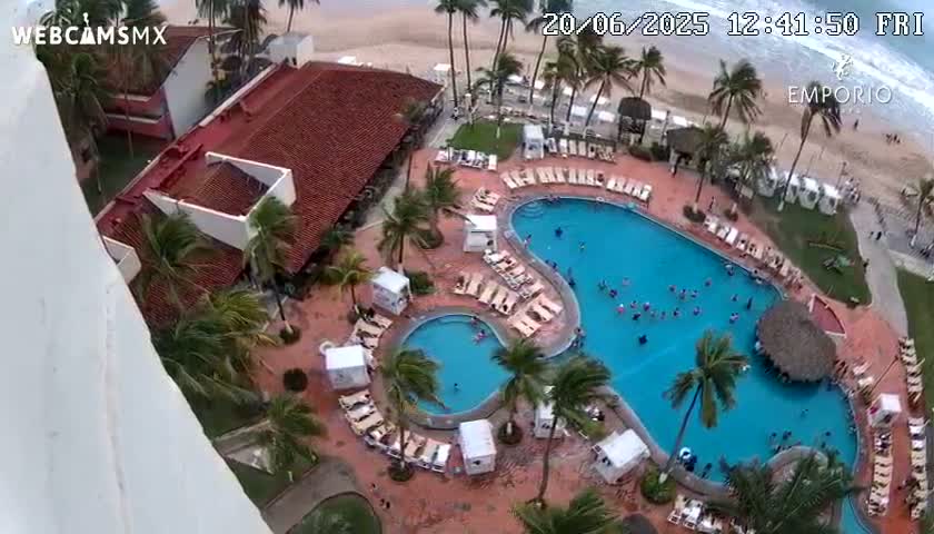 An aerial view shows a resort pool complex with many people swimming, surrounded by palm trees, buildings, and a beach with ocean waves visible in the background under a windy sky.