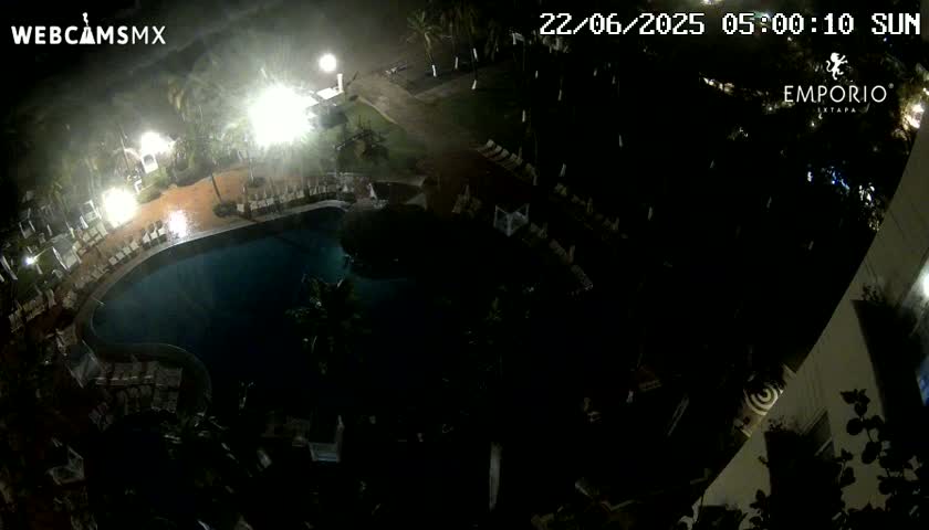A nighttime overhead view of a resort pool area during a rain shower, showing lounge chairs surrounding the pool and palm trees in the surrounding area.