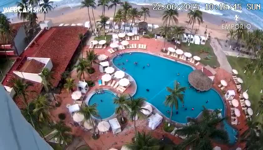 An aerial view shows a resort pool area with many people swimming and lounging on sunny day, next to an ocean with waves.