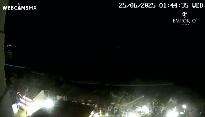 A nighttime, high-angle view shows a dark ocean, a dimly lit beach area with buildings and vegetation, and some bright lights.