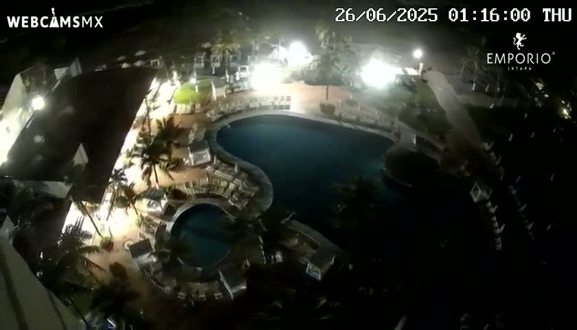 An aerial nighttime view shows a resort pool area with palm trees and surrounding buildings under bright artificial lighting.