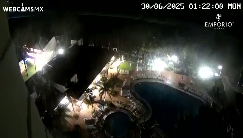 A nighttime, high-angle view shows a resort complex with pools and palm trees illuminated by artificial lights under a dark, possibly rainy sky.