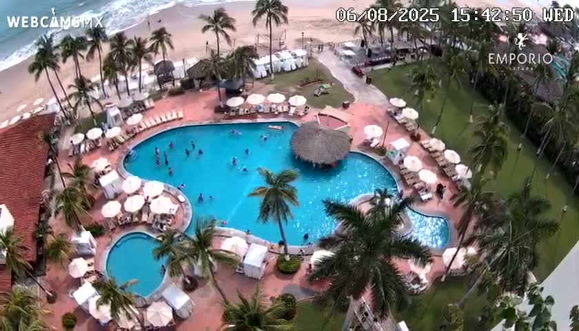 An aerial view shows a resort pool area with multiple pools, palm trees, lounge chairs with umbrellas, and an ocean beach in the background on a partly sunny day.