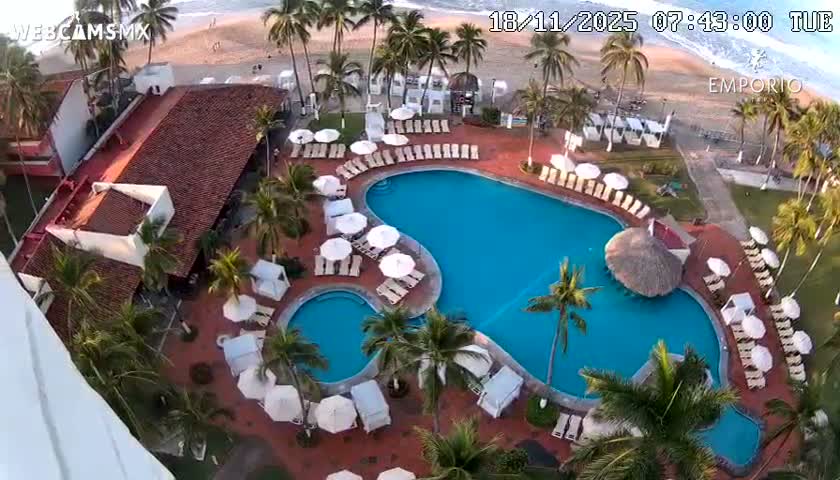 An aerial view shows a resort pool area with multiple pools, palm trees, lounge chairs with umbrellas, and an ocean beach in the background on a partly sunny day.