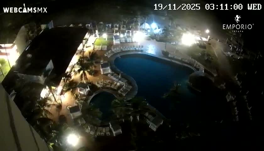 An aerial night view reveals two illuminated swimming pools surrounded by lounge chairs, cabanas, and palm trees at a resort, under clear weather conditions.