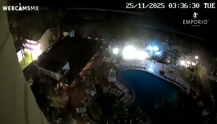 An aerial view captures a dimly lit resort scene at night, showcasing a dark blue swimming pool, palm trees, and various brightly illuminated outdoor dining and lounge areas stretching towards the dark ocean, under clear weather.
