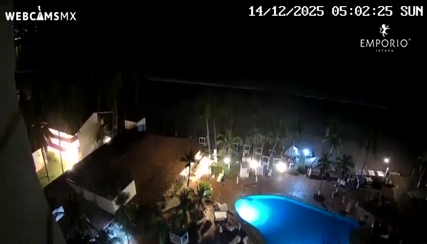 An aerial view captures a dimly lit resort scene at night, showcasing a dark blue swimming pool, palm trees, and various brightly illuminated outdoor dining and lounge areas stretching towards the dark ocean, under clear weather.