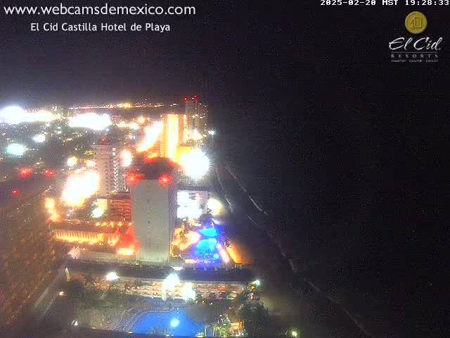 A brightly lit resort complex at night stretches along a dark beach under a clear sky.