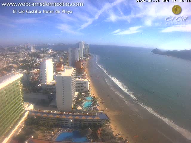 A high-angle, sunny view shows a long sandy beach meeting a calm ocean next to a city with many high-rise hotels.