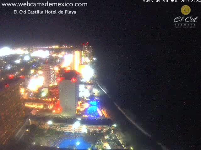 A brightly lit resort town at night, seen from above, extends to a dark ocean under a clear, starlit sky.