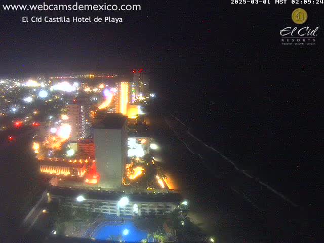 A nighttime aerial view of a brightly lit coastal city with a dark ocean and calm waves under a clear sky.