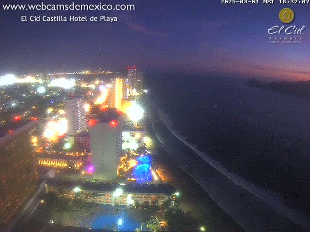 An aerial nighttime view of a brightly lit coastal city with a beach, showing the ocean under a twilight sky.