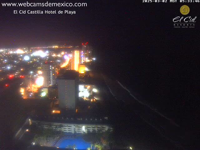 A nighttime aerial view shows a brightly lit coastal city with a dark ocean and beach stretching along the bottom.