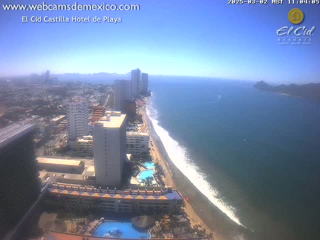 A high-angle, sunny-day view shows a long sandy beach with gentle waves lapping the shore, lined with hotels and high-rise buildings, extending into a city landscape.