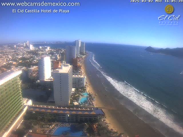 An aerial view shows a sandy beach with gentle waves lapping the shore, under a clear blue sky, next to a city with many high-rise hotels and buildings.