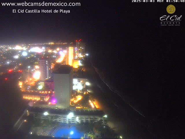 A nighttime aerial view shows brightly lit hotels and city lights along a dark coastline under a clear sky.