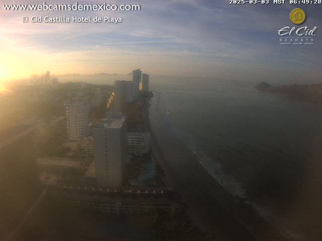 A hazy sunrise illuminates a coastal city with high-rise buildings and a dark-sand beach next to a calm ocean.