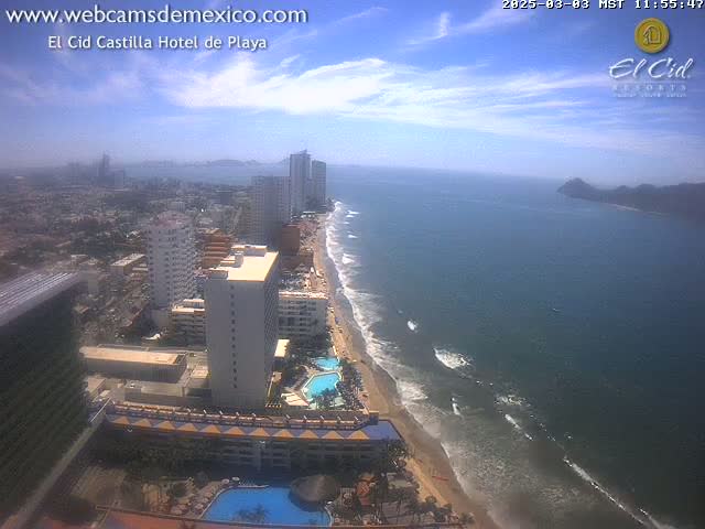 An aerial view shows a long sandy beach with gentle waves, numerous high-rise hotels, and a partly cloudy sky.
