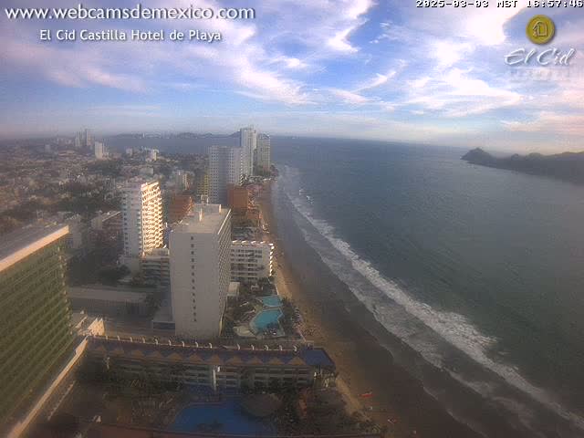 An aerial view shows a beach with gentle waves, numerous high-rise hotels, and a partly cloudy sky.