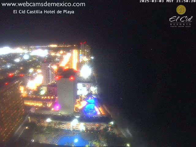A brightly lit resort area at night, seen from above, extends toward a dark ocean under a clear night sky.