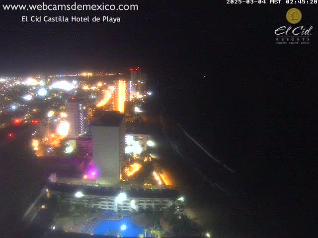 A nighttime aerial view shows a brightly lit city coastline with high-rise hotels and a swimming pool, extending to a dark ocean under a clear sky.