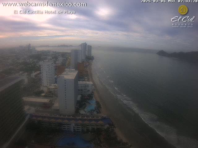 An aerial view shows a beach with gentle waves, a coastline of hotels and buildings under a hazy, overcast sky.