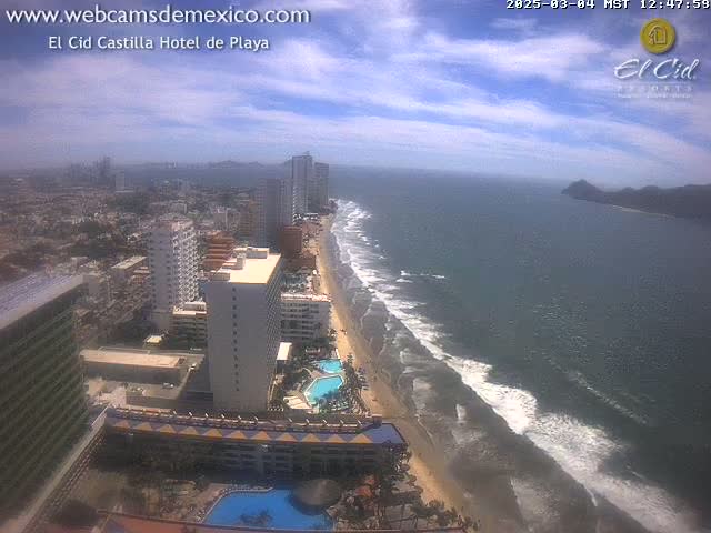 An aerial view shows a beach with waves under a partly cloudy sky, next to a city with high-rise hotels and buildings.