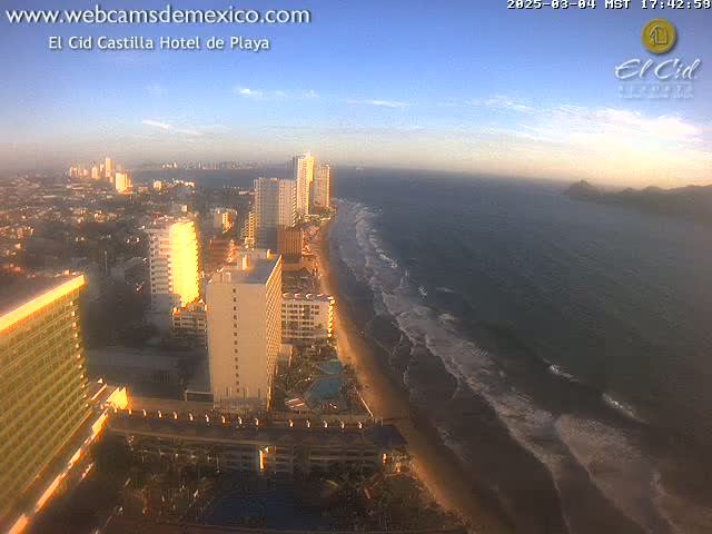 An aerial view of a beach city at sunset, showing high-rise hotels along a sandy beach with gentle waves lapping the shore.