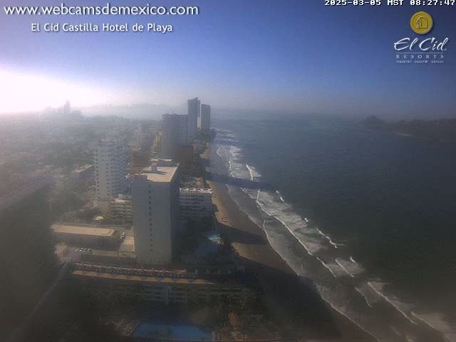 A hazy, sunny day shows a beach with waves lapping a sandy shore, lined with tall buildings and hotels, stretching into a city in the distance.