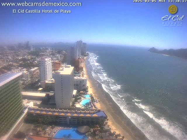 An aerial view shows a beach with gentle waves under a clear, sunny sky, next to a city with many high-rise hotels and buildings.