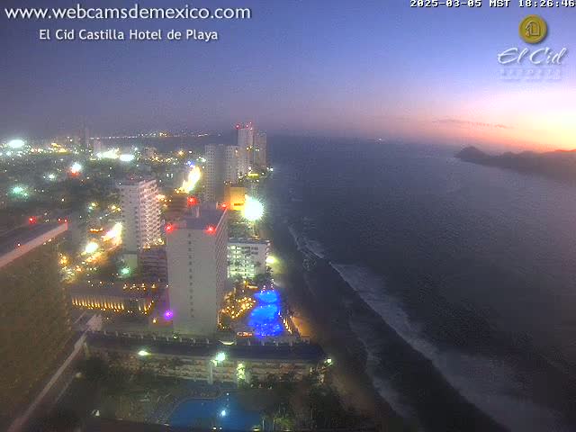 An aerial night view of a coastal city with brightly lit high-rise hotels and buildings along a dark sandy beach under a twilight sky.