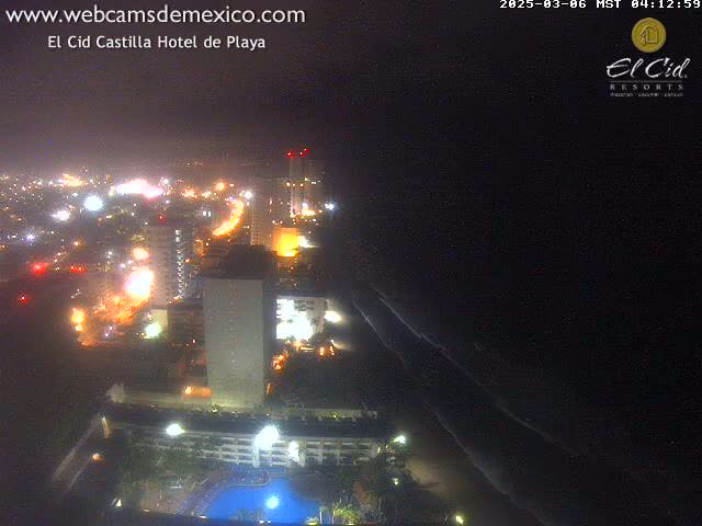 A nighttime, hazy view of a city illuminated by lights along a coastline with dark ocean waves.