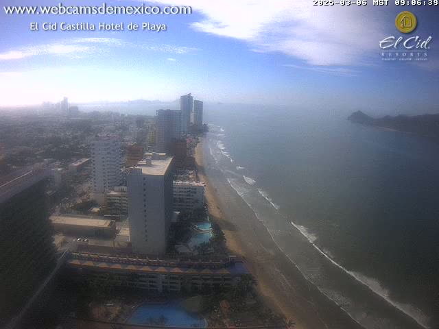 An aerial view of a hazy beach, ocean, and cityscape with high-rise hotels under a partly cloudy sky.
