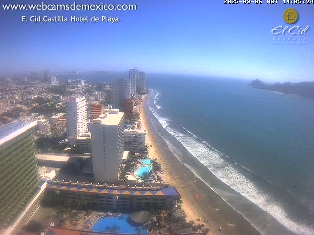 An aerial view shows a long sandy beach with gentle waves under a clear blue sky, backed by numerous high-rise hotels and a sprawling city.