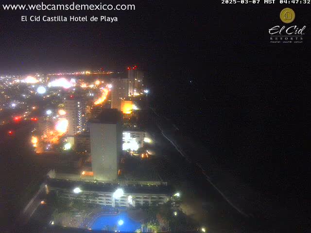 A nighttime, aerial view shows a brightly lit coastal city next to a dark ocean, with a large hotel and swimming pool visible in the foreground.