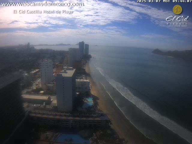 An aerial view shows a beach with waves lapping the shore, lined with high-rise hotels and buildings under a partly cloudy sky.