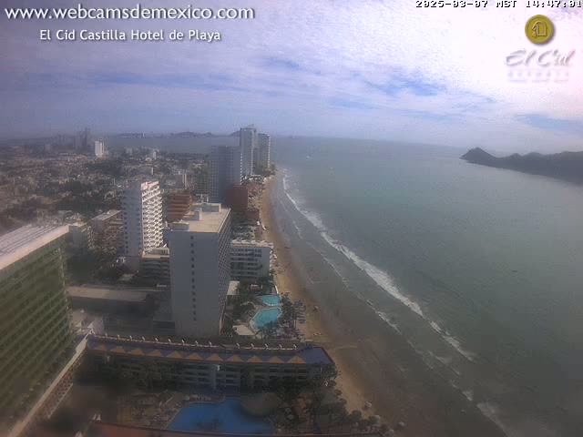 A long sandy beach stretches alongside a calm ocean, lined with numerous high-rise hotels and buildings under a partly cloudy sky.