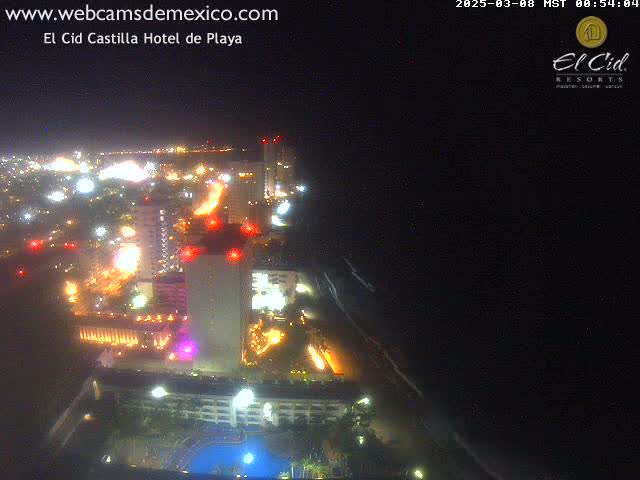A nighttime aerial view of a brightly lit coastal city with hotels and a beach, under a clear, dark sky.
